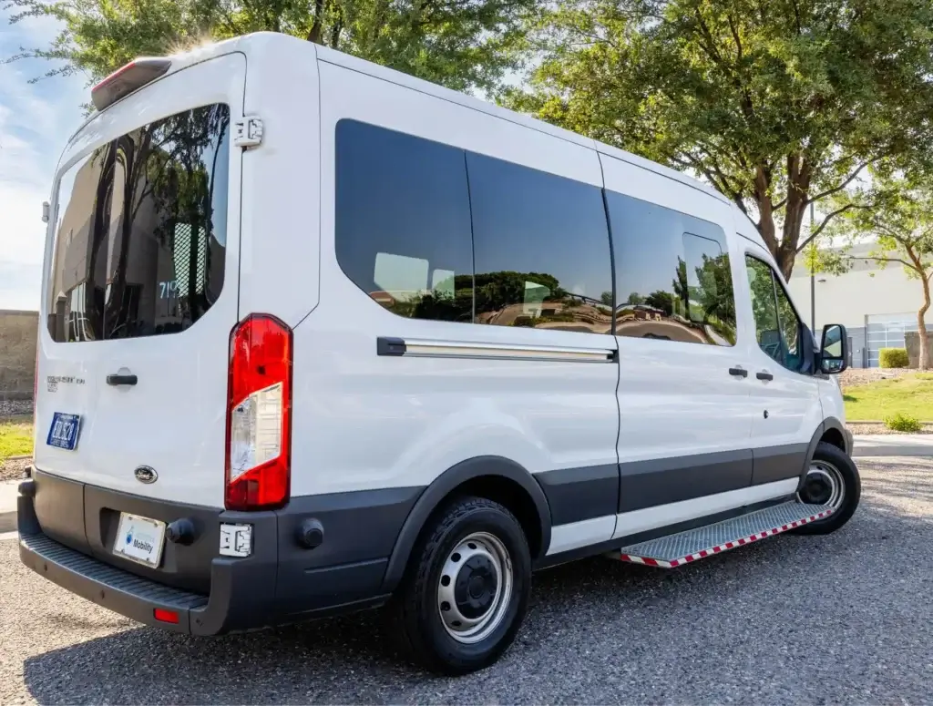 A white passenger van is parked on a paved area, with trees and buildings visible in the background.