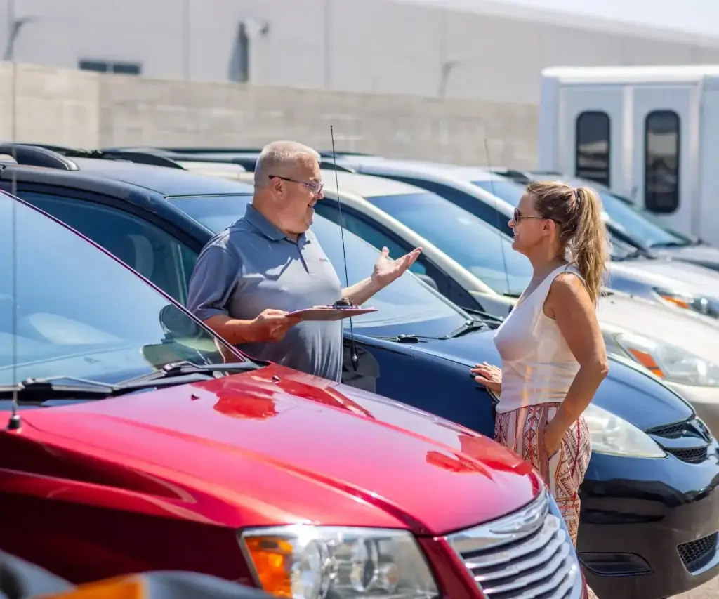 A man and a woman conversing in front of a row of parked cars at a dealership. The man is holding a clipboard.
