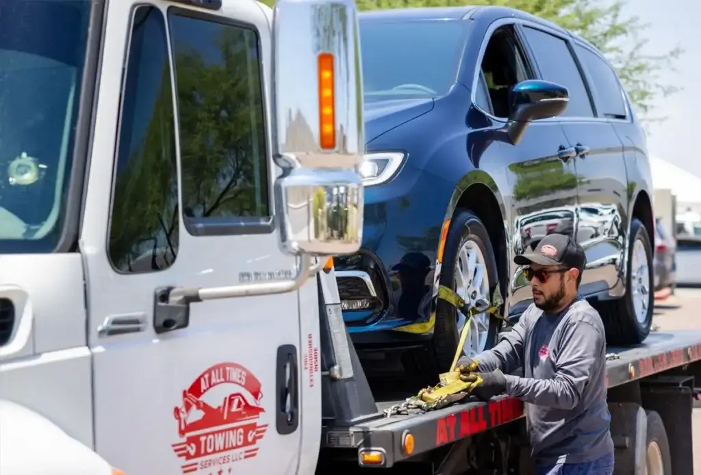 Tow truck driver secures a blue minivan on a flatbed truck using a chain.