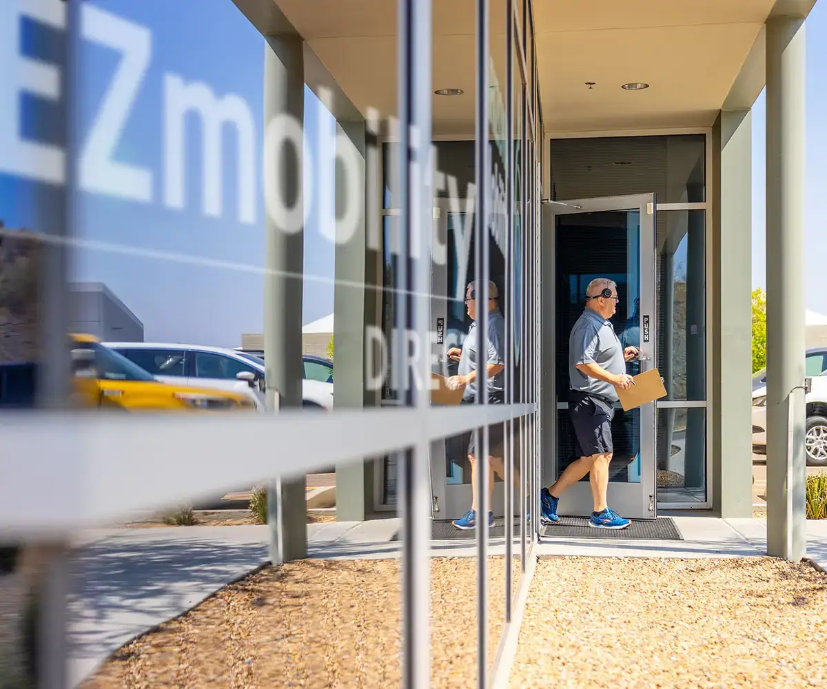 Person carrying a package enters a glass-fronted building labeled EZmobility, with reflections of cars and outdoor scenery visible.