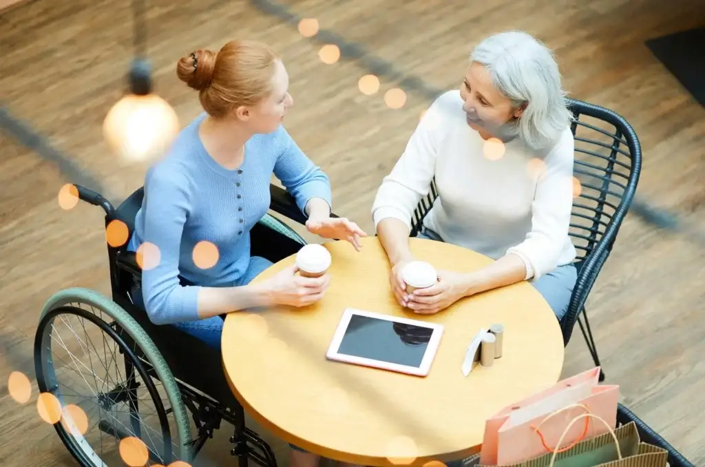 Two women sit at a round table with coffee cups, a tablet, and shopping bags. One woman is in a wheelchair. They appear to be engaged in conversation.
