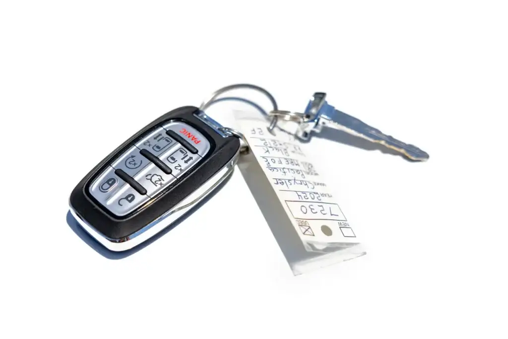 A car key fob with buttons for lock, unlock, and alarm features, attached to a key and a white tag with handwritten numbers.