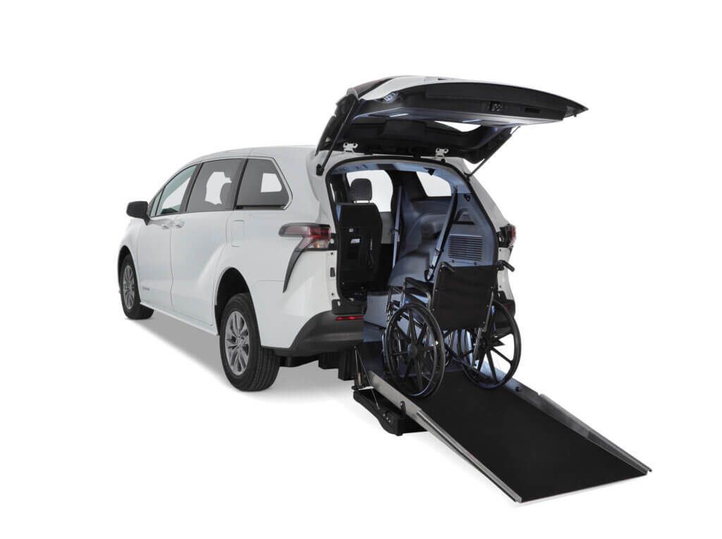 A white minivan with its rear hatch open features a deployed ramp and a wheelchair positioned inside the vehicle.