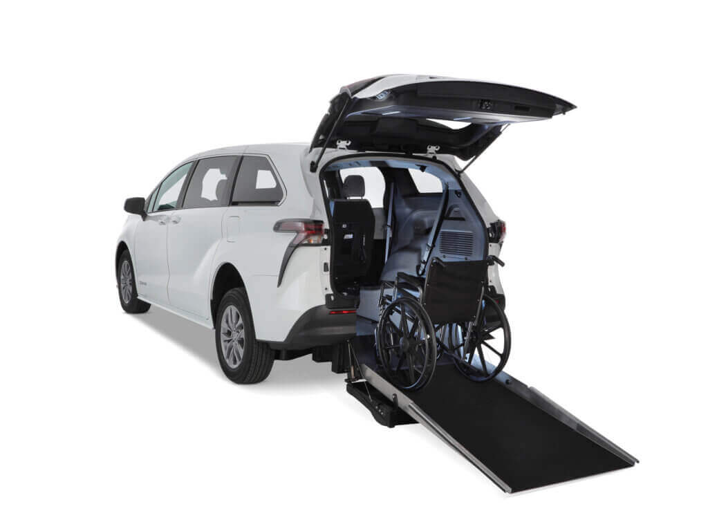A white minivan with its rear hatch open features a deployed ramp and a wheelchair positioned inside the vehicle.